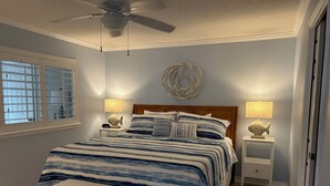 Een slaapkamer met een bed, nachtkastjes, een plafondventilator, decoratie aan de muur en een raam met jaloezieën.