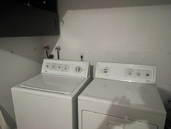 Two white front-loading washing machines in a laundry room with a white wall and a dark shelf above.