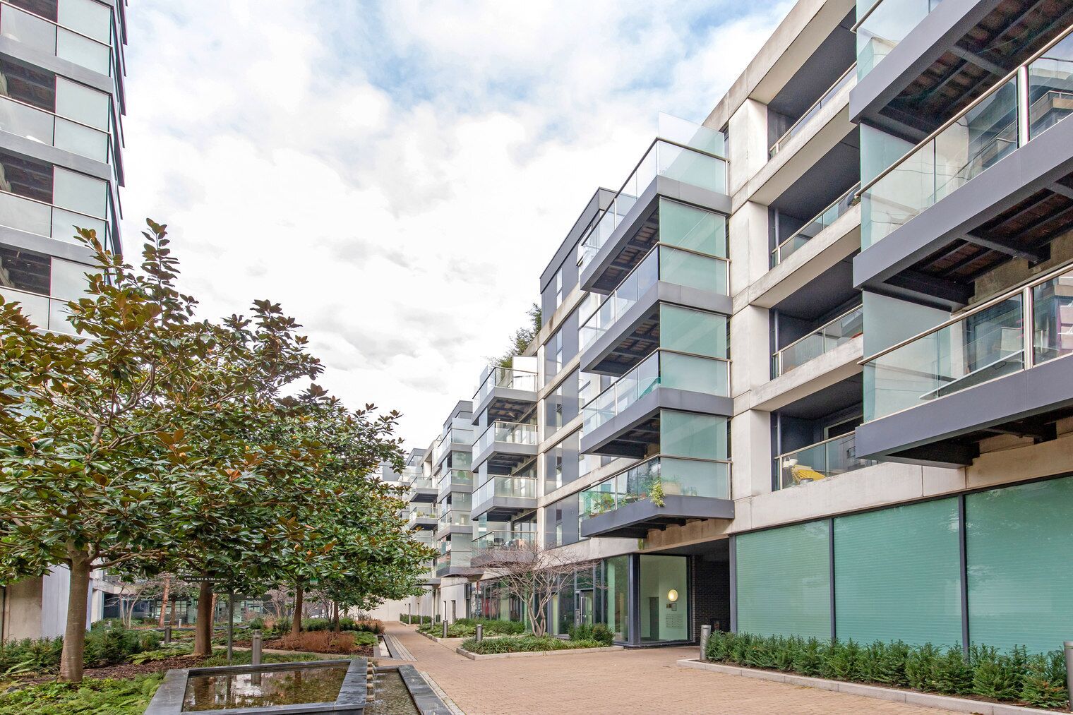 A modern multi-story residential building with balconies, a paved walkway, and greenery.