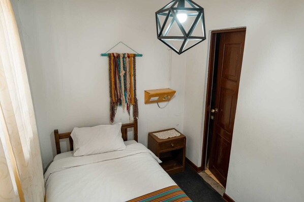 Un dormitorio pequeño y ordenado con cama, mesita de noche, un tapiz colgante y una lámpara de techo geométrico.