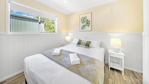 A hotel room with a large bed, two bedside tables with lamps, a window with blinds, and a framed plant on the wall. - Hideaway Mission Beach Village (Mission Beach)