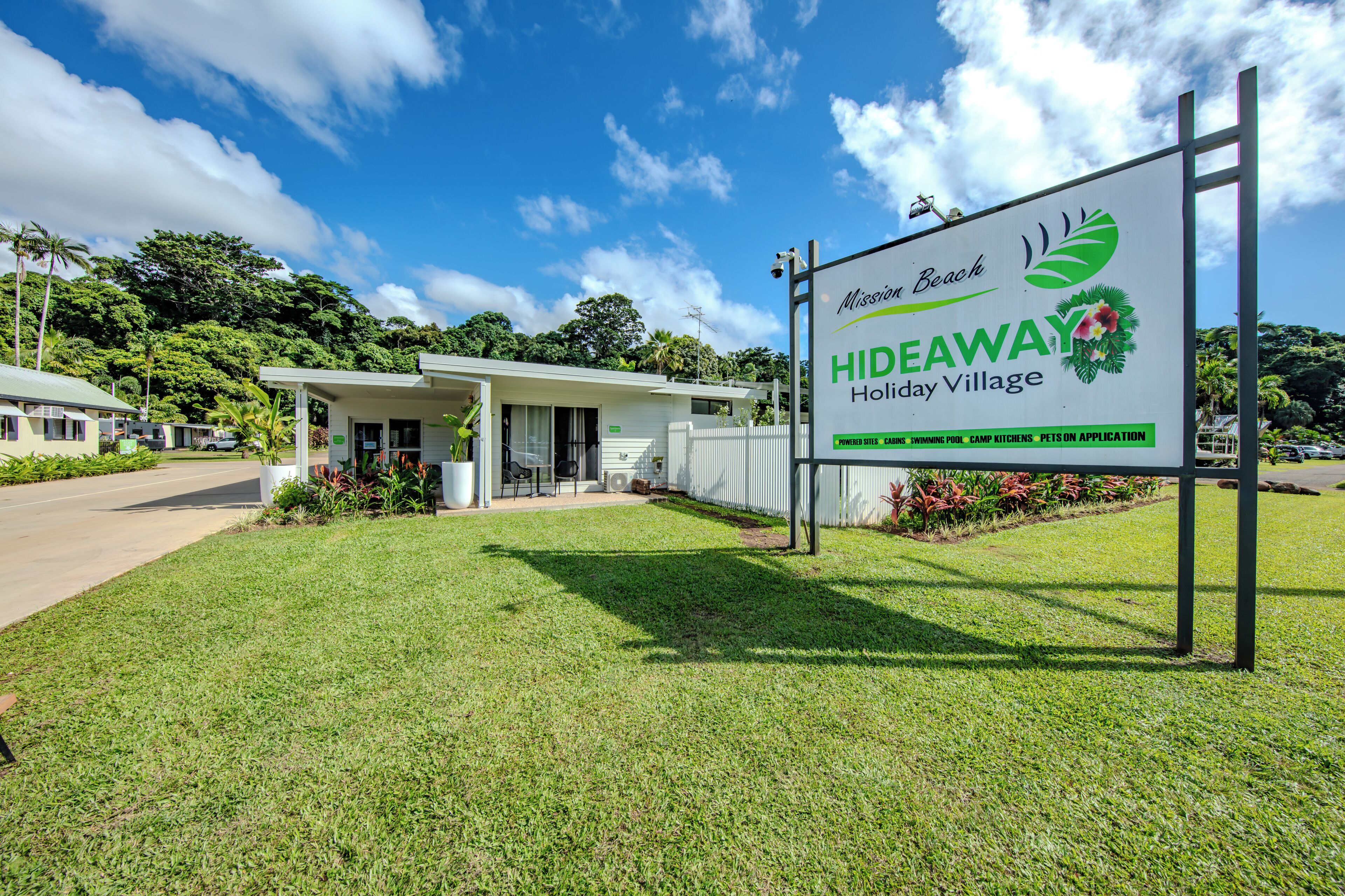 A sign for Mission Beach Hideaway Holiday Village with a white building and a grassy area in front.