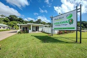 A sign for Mission Beach Hideaway Holiday Village with a white building and a grassy area in front.