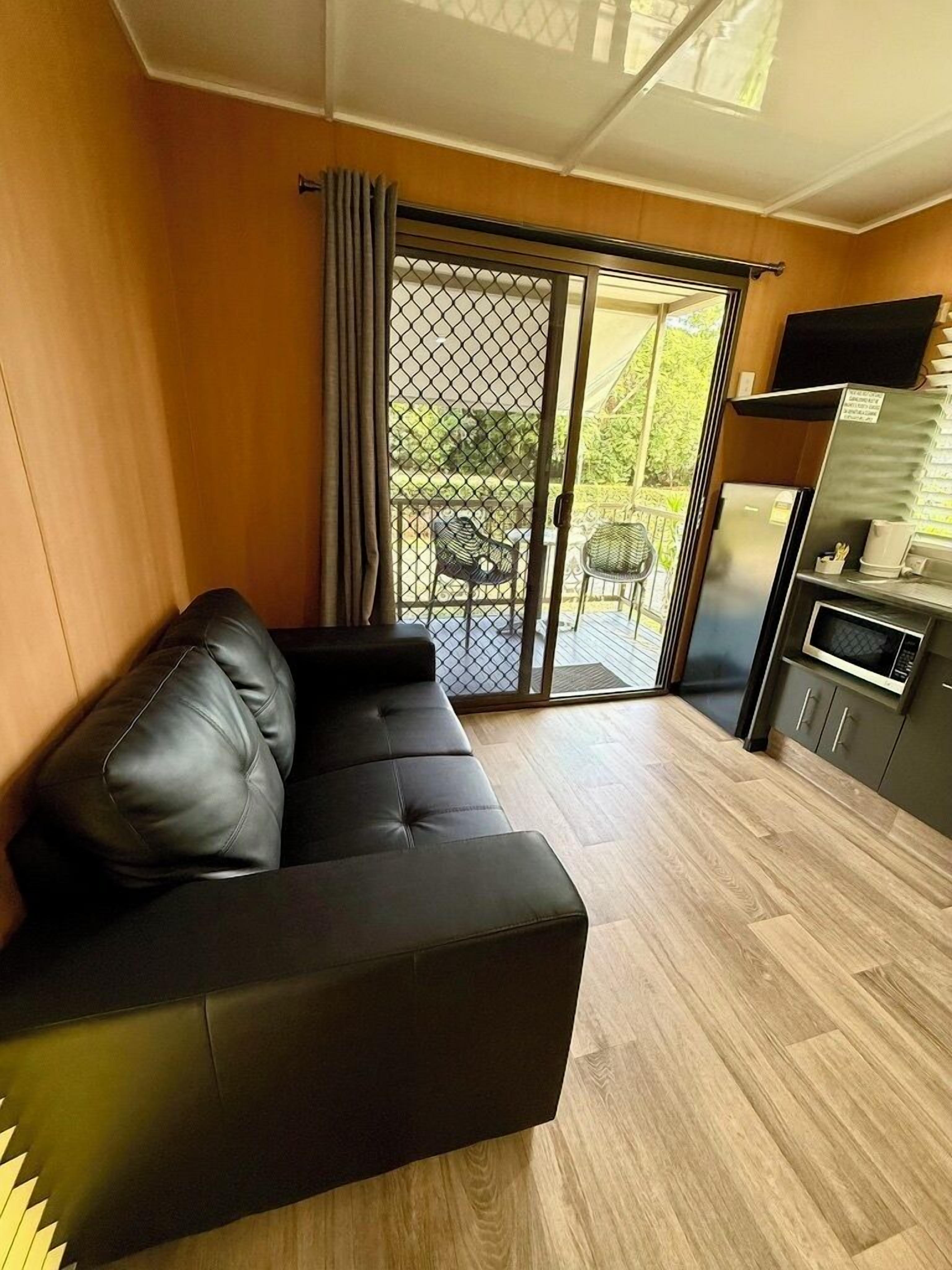 A modern living room with a black leather sofa, a wooden floor, a sliding glass door leading to an outdoor area with a table and chairs, and a kitchen area with a refrigerator and microwave.
