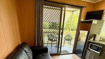 A modern living room with a black leather sofa, a wooden floor, a sliding glass door leading to an outdoor area with a table and chairs, and a kitchen area with a refrigerator and microwave.