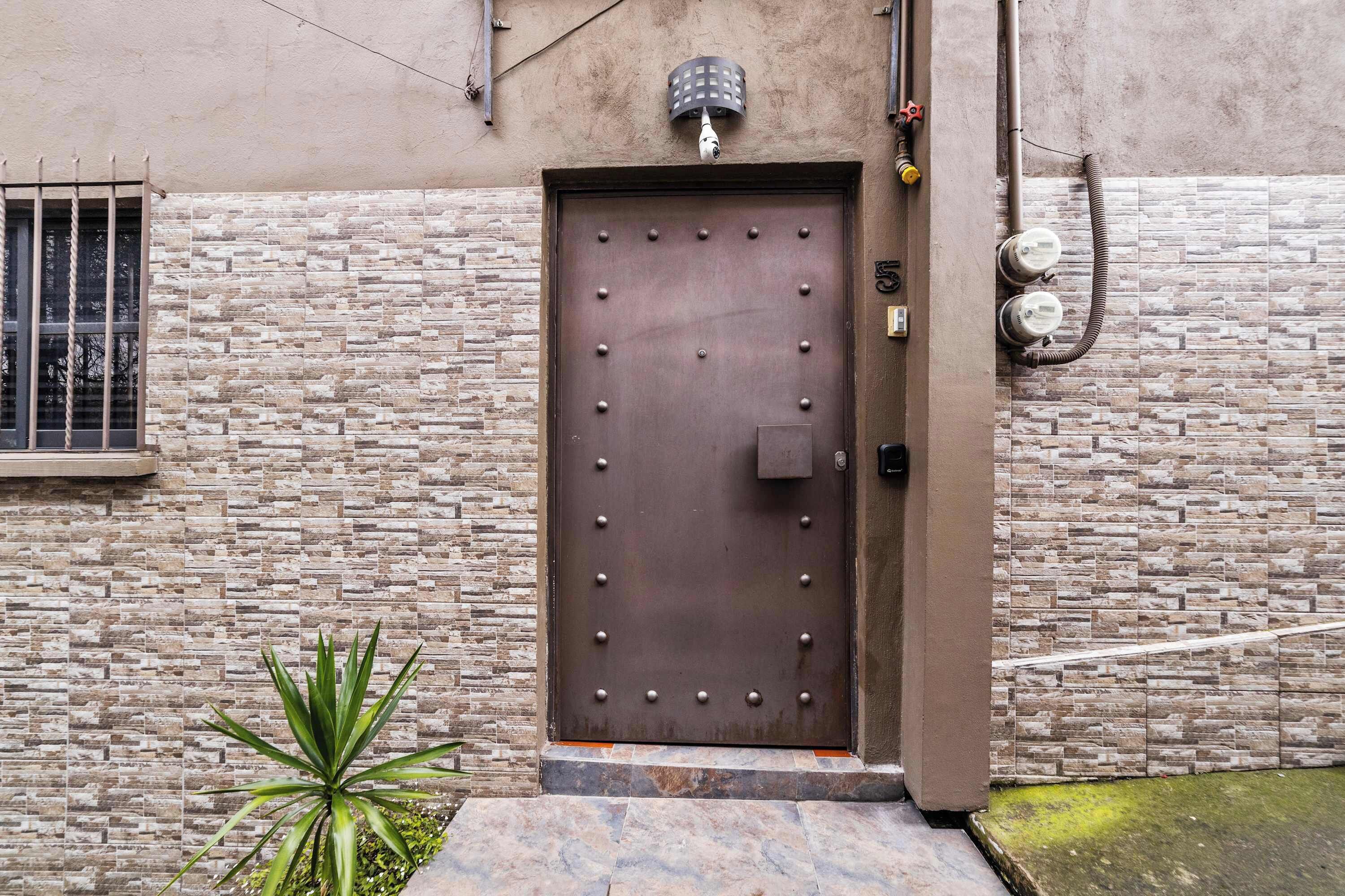 Una puerta marrón con remaches de metal, un pequeño ventanal con barrotes y una planta en maceta a su lado.