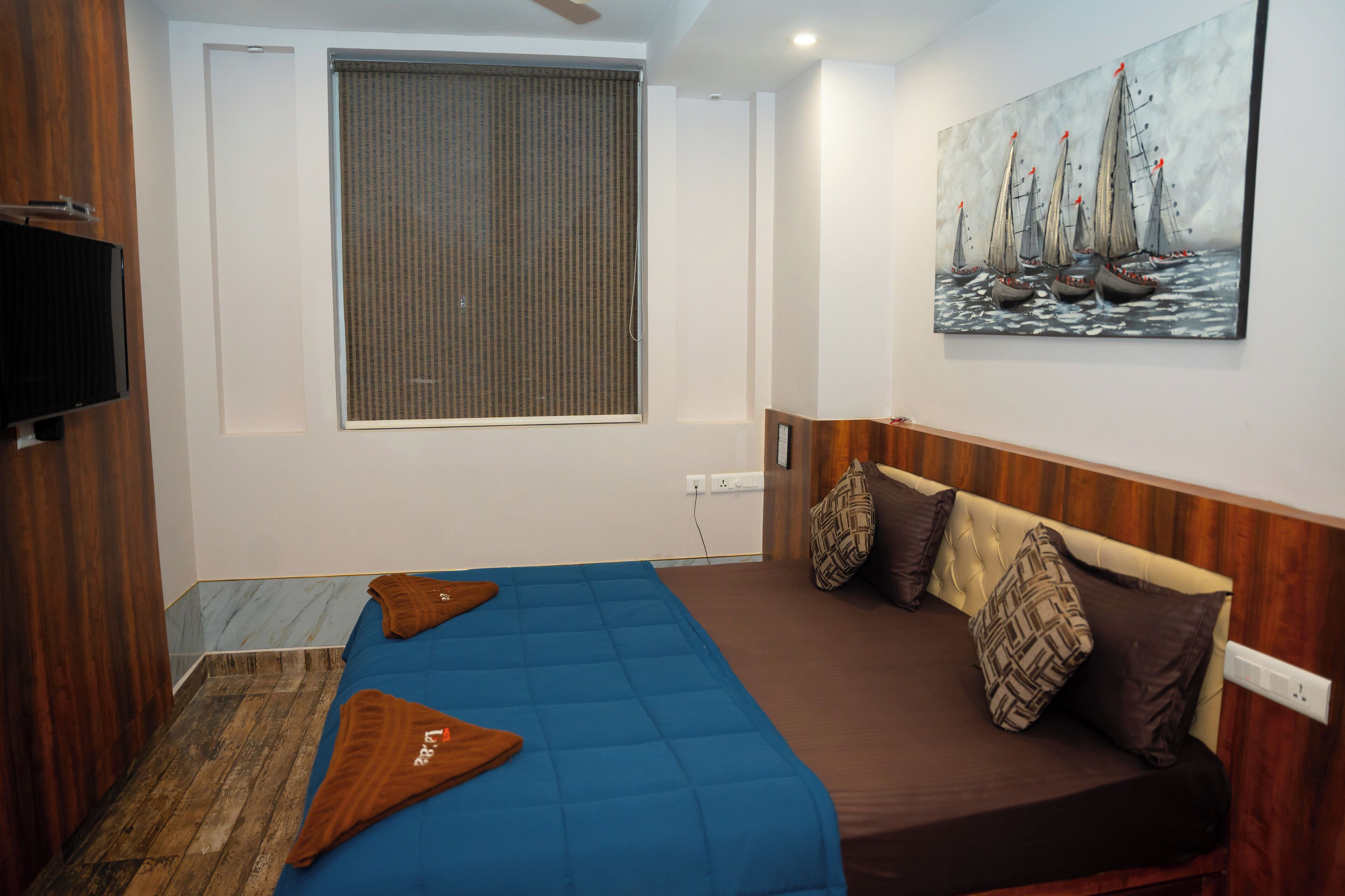 A hotel room with a bed, a TV, a painting, and a window with blinds.