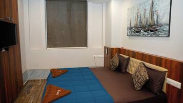 A hotel room with a bed, a TV, a painting, and a window with blinds.