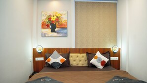 Une chambre d’hôtel avec un lit, un tableau représentant des fleurs, une tête de lit agrémentée de coussins décoratifs et une prise électrique au mur.