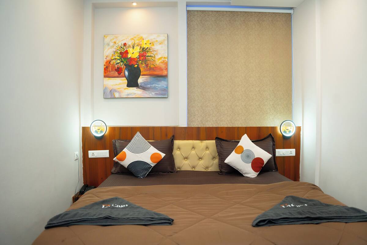 A hotel room with a bed, a painting of flowers, a headboard with decorative pillows, and a wall socket.
