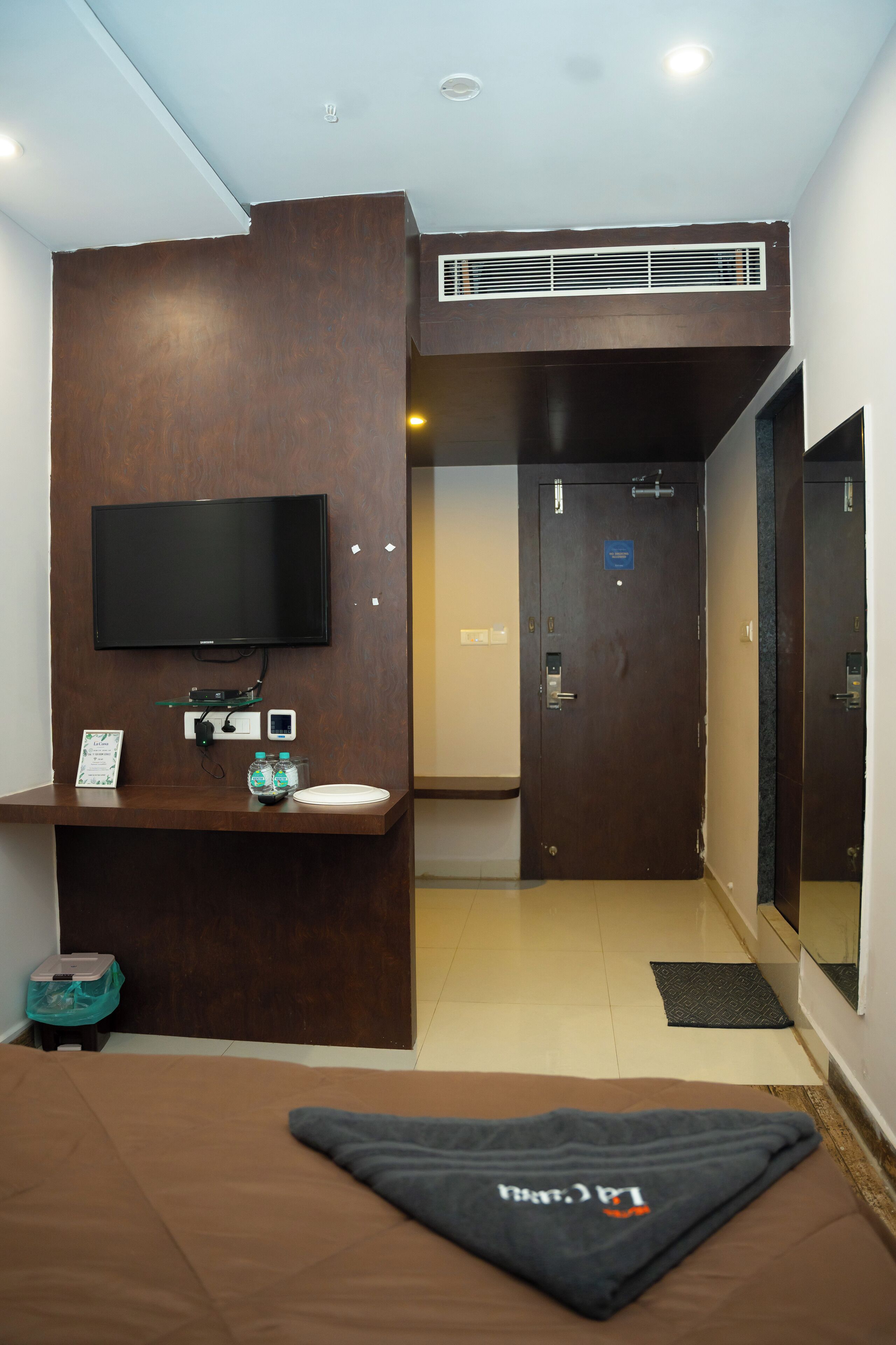 A compact hotel room with a bed, a TV mounted on the wall, a small desk with a computer and a bottle, and a door leading to another room.
