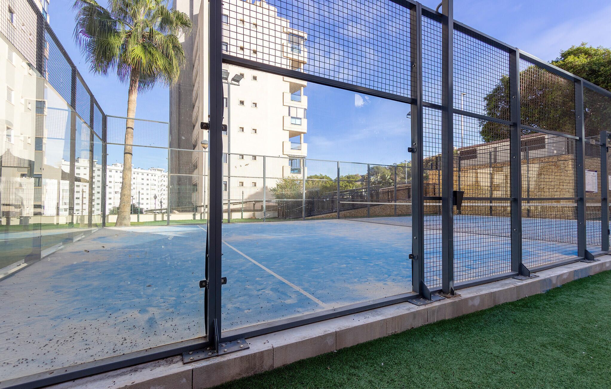 An indoor tennis court with a net, surrounded by a metal fence, and adjacent to a building with multiple windows and balconies.
