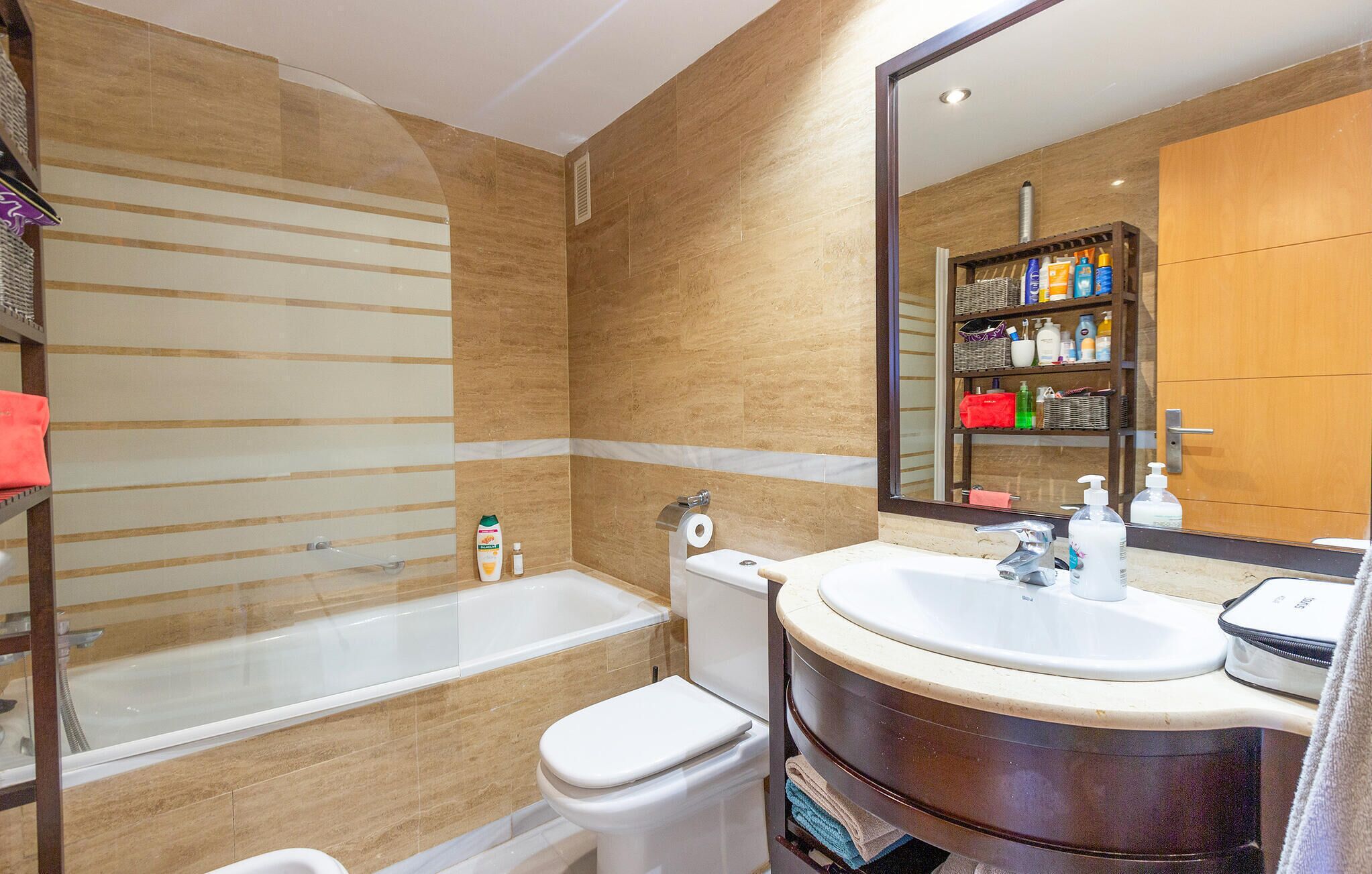 Ein modernes Badezimmer mit Badewanne, Toilette und Waschbecken. Über dem Waschbecken hängt ein Spiegel, darüber befindet sich ein Ablagefach mit Toilettenartikeln.