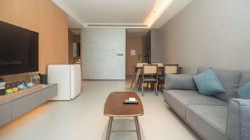 A modern living room with a flat-screen TV, a white refrigerator, a wooden coffee table, and a grey sofa with blue cushions.