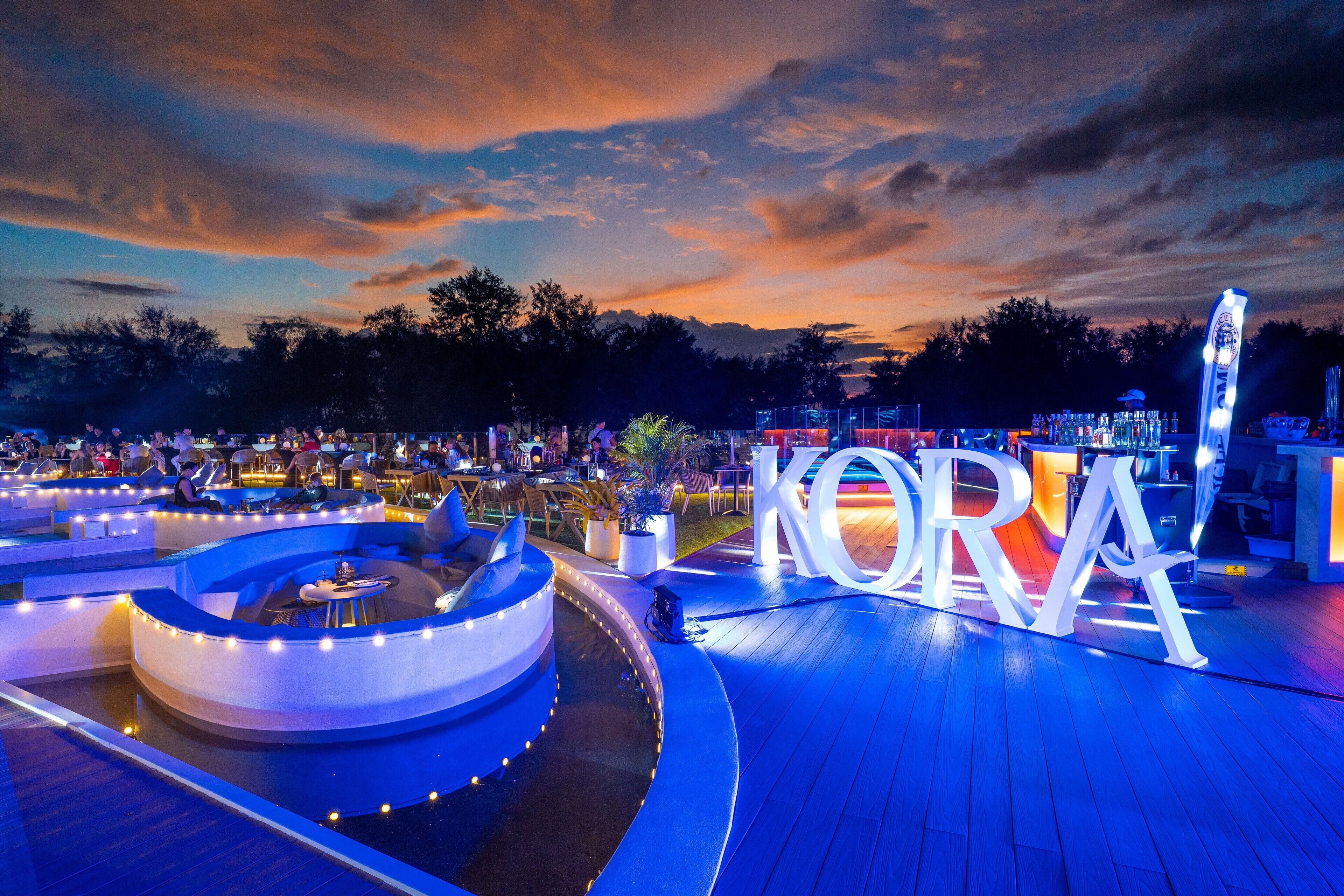 A waterfront hotel with a large illuminated sign reading "KORVA," a circular water feature, and outdoor seating areas.