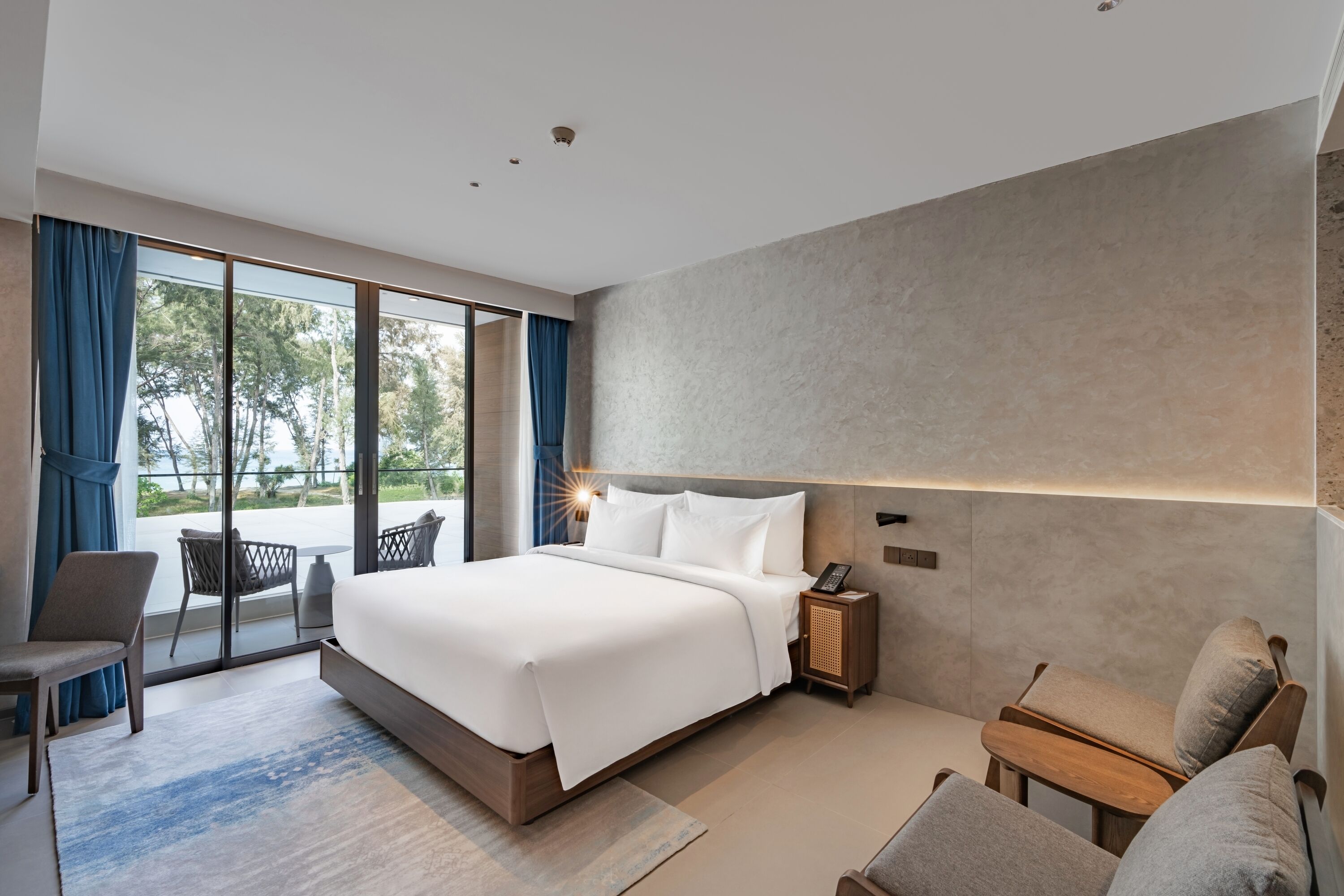 Una habitación de hotel moderna con una cama grande, un área de descanso con una silla y una mesita, una mesita de noche con lámpara, y vistas a través de la puerta de vidrio.