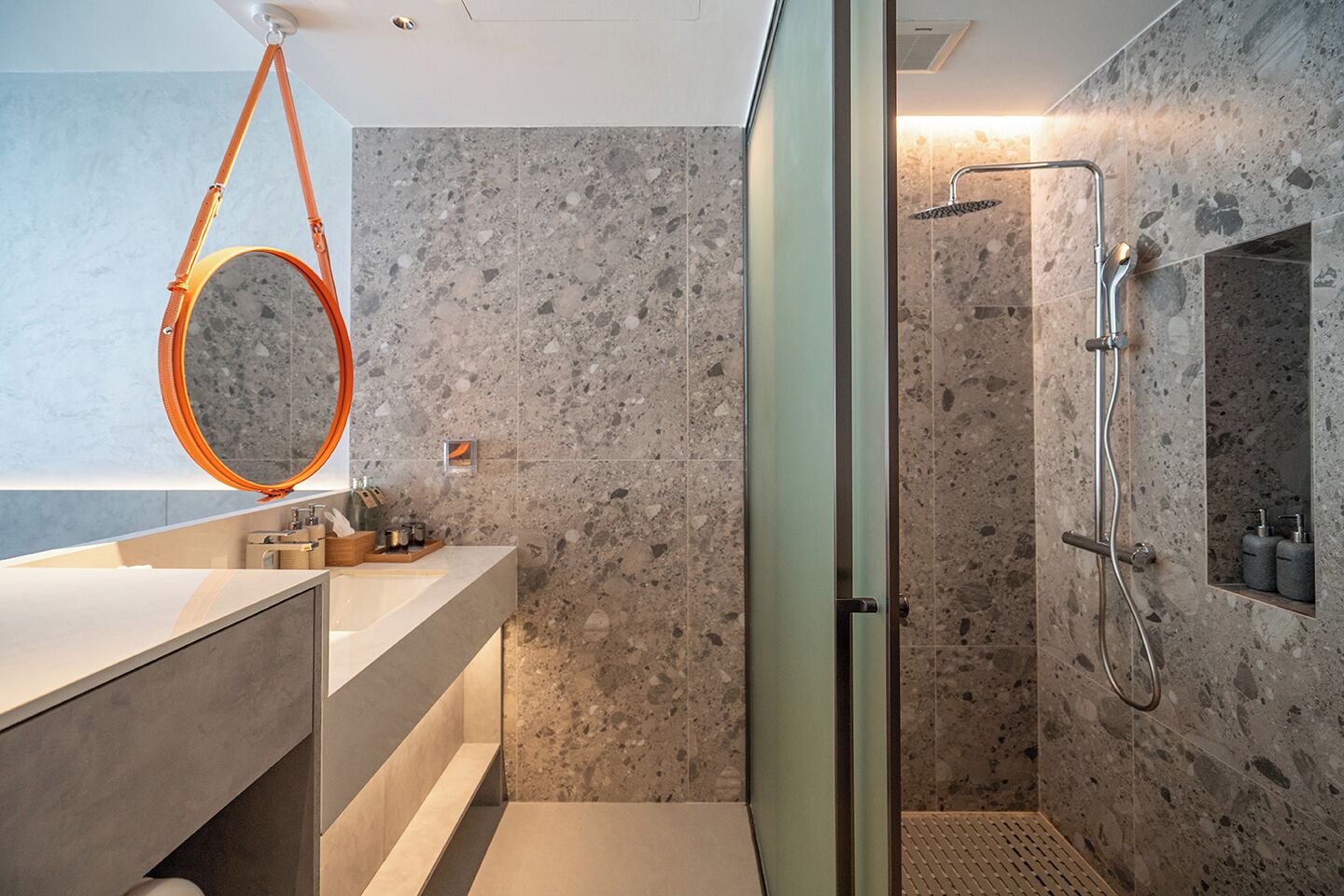 A modern bathroom with a glass shower enclosure, a marble-tiled wall, and a round mirror with an orange frame.