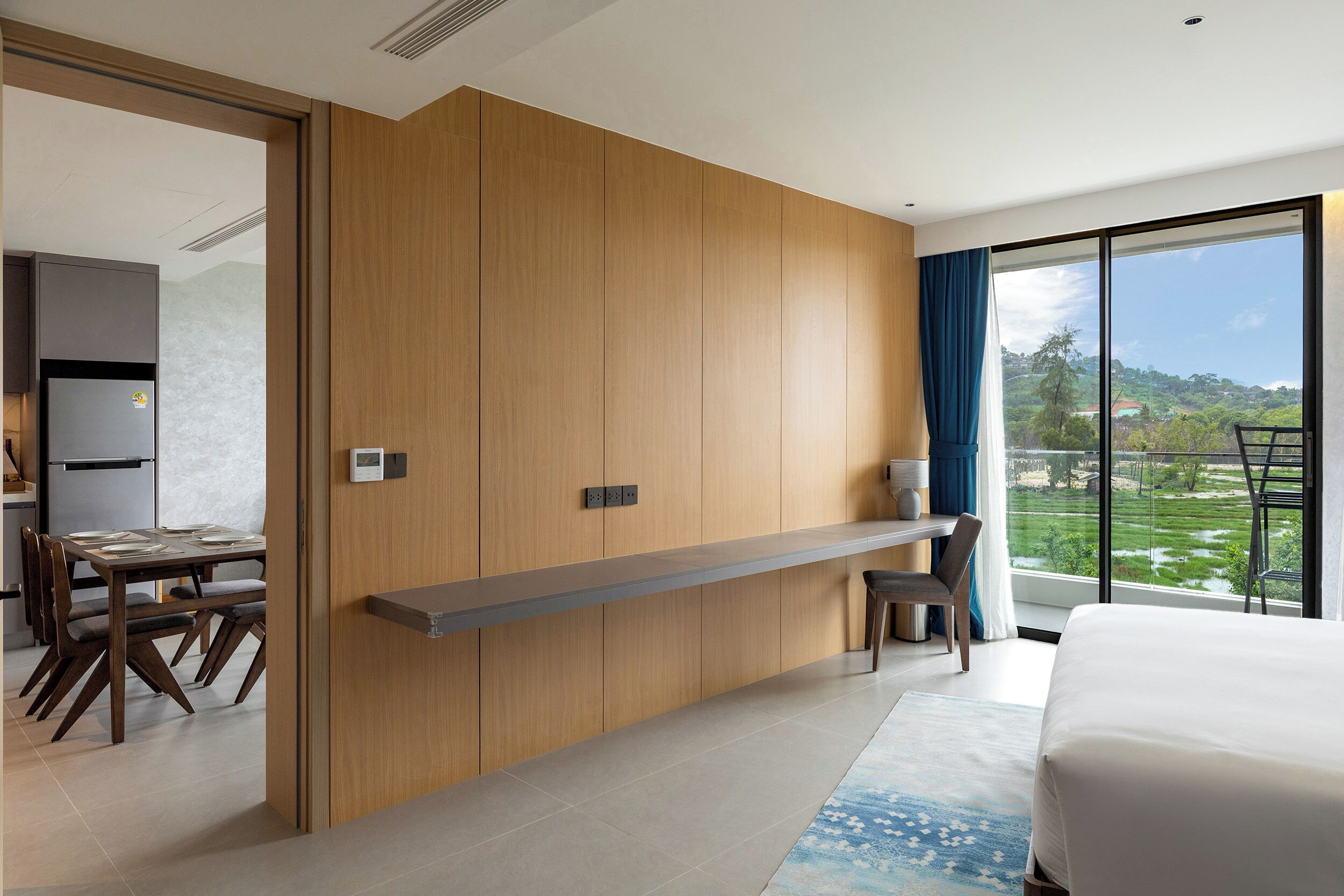 A modern hotel room with a large bed, a dining area with a table and chairs, a built-in bench, and a large window with a view of the landscape.