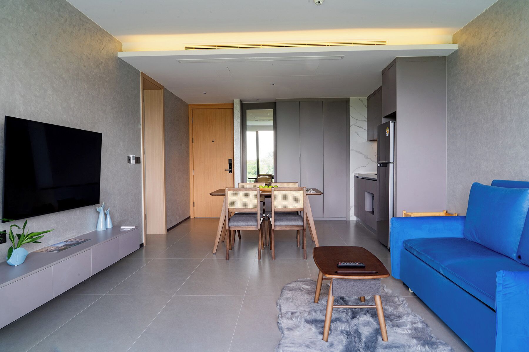 Una sala de estar moderna con televisor de pantalla plana, un sofá azul, un área de comedor con mesa y sillas, y una cocina con refrigerador.