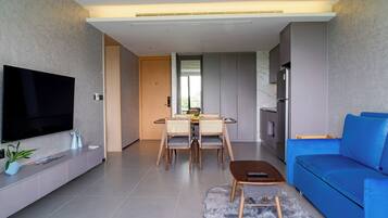 Una sala de estar moderna con televisor de pantalla plana, un sofá azul, un área de comedor con mesa y sillas, y una cocina con refrigerador.