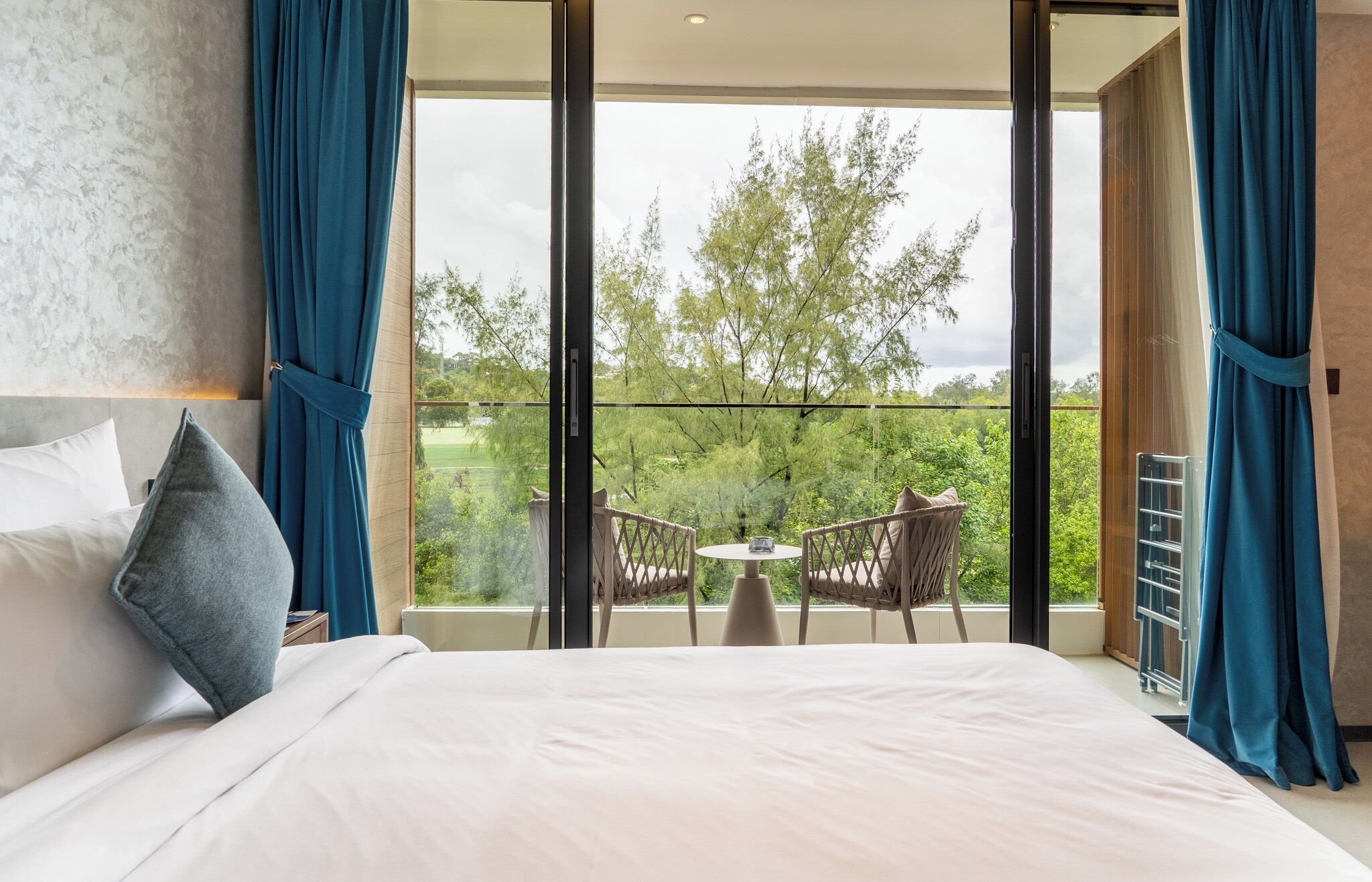A hotel room with a large bed, a view of trees through floor-to-ceiling windows, and a small balcony with a table and chairs.