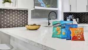 A kitchen with white cabinets, a marble countertop, and a window with blinds. There are bags of snacks on the counter.