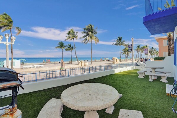 A beachfront patio with a round table and benches, palm trees, and a view of the ocean.
