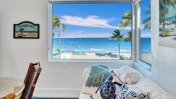 A room with a large window overlooking a beach, a framed sign on the wall, and a chair with a decorative backrest. - Sunset Oceanfront Villa (Hollywood)