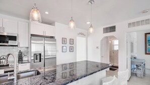 A modern kitchen with white cabinetry, a granite countertop, stainless steel appliances, pendant lighting, and a view into the dining area. - Sunset Oceanfront Villa (Hollywood)