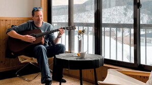Une personne joue de la guitare dans une pièce dotée d’une grande fenêtre, d’une petite table sur laquelle se trouvent une tasse et des fleurs, ainsi que d’une chaise.