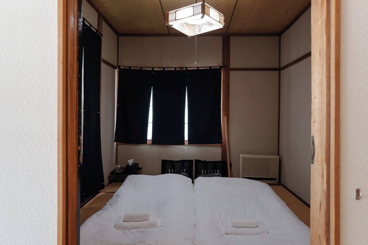 A traditional Japanese-style room with tatami flooring, a futon bed, a sliding door, and a ceiling light fixture.