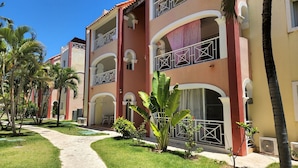 A hotel with a red and beige facade, multiple balconies, and a pathway lined with palm trees. - Bavaro-El Cortecito Beach Cozy apartment with AC and Fast WiFi. No cleaning fee (Punta Cana)