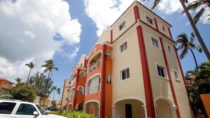 A multi-story hotel with balconies and arched windows, surrounded by palm trees and parked cars. - Bavaro-El Cortecito Beach Cozy apartment with AC and Fast WiFi. No cleaning fee (Punta Cana)