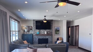 A modern living room with a fireplace, a dining table with chairs, a ceiling fan, and a large TV mounted on the wall.
