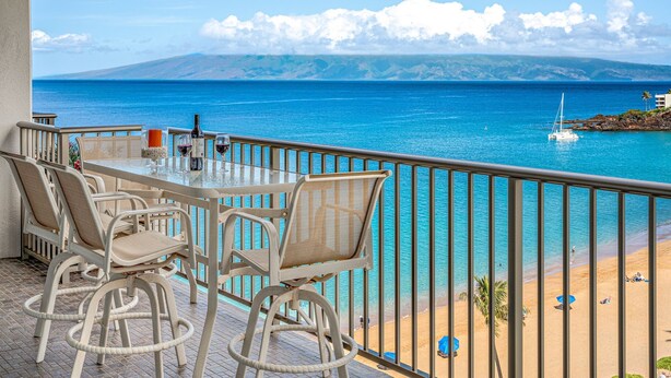 A balcony with wicker chairs and a table, overlooking a beach and the sea.