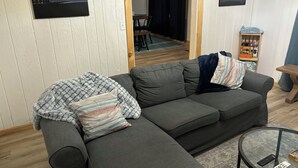 Un canapé d’angle gris avec des coussins à rayures et unis, une table basse en verre avec des télécommandes, et deux tableaux encadrés accrochés au mur.