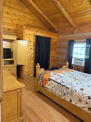 A wooden cabin room with a bed, a television, a wardrobe, and a window with curtains.