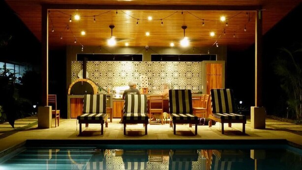 Un área junto a la piscina con una terraza de madera, una zona de bar con un revestimiento estampado y varias tumbonas a rayas.