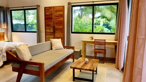 Una habitación acogedora con un sofá de madera, una pequeña mesa de madera y un escritorio con una silla. Amplias ventanas ofrecen vistas al verdor exterior.