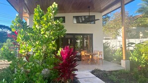 Un patio cubierto con una mesa y sillas para comer, rodeado de follaje y la entrada de un edificio.
