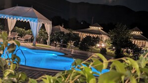 A nighttime view of a pool area with a tent structure, surrounded by lush greenery and a mountainous backdrop. - Arvana Resort & Spa Pushkar (Pushkar)