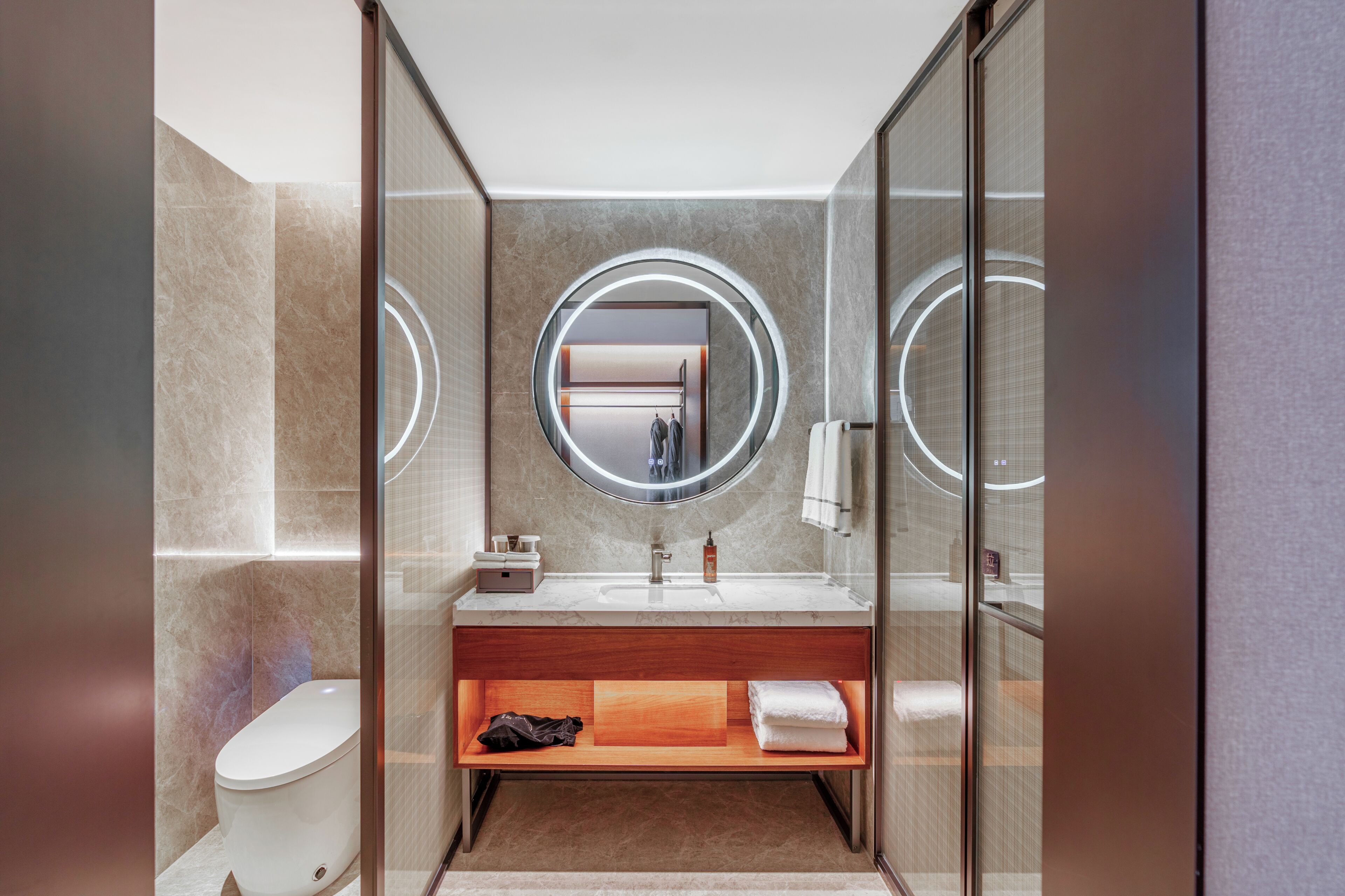 A modern bathroom with a marble vanity, a round mirror, a toilet, and a towel rack.