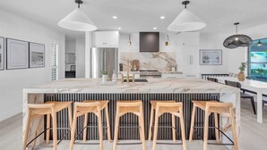 Una cocina moderna con una isla de mármol, taburetes de bar, iluminación colgante y una mesa de comedor grande.