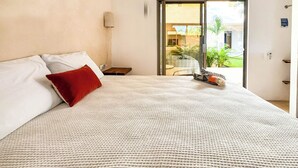 A bedroom with a bed, a red pillow, a nightstand, a lamp, and a sliding glass door leading to a balcony with plants. - Aria (El Cuyo)