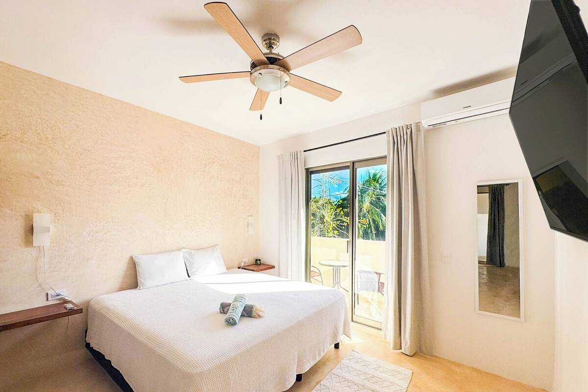 Une chambre d’hôtel équipée d’un lit, d’un ventilateur de plafond, d’une télévision et d’une porte-fenêtre coulissante donnant sur l’extérieur.