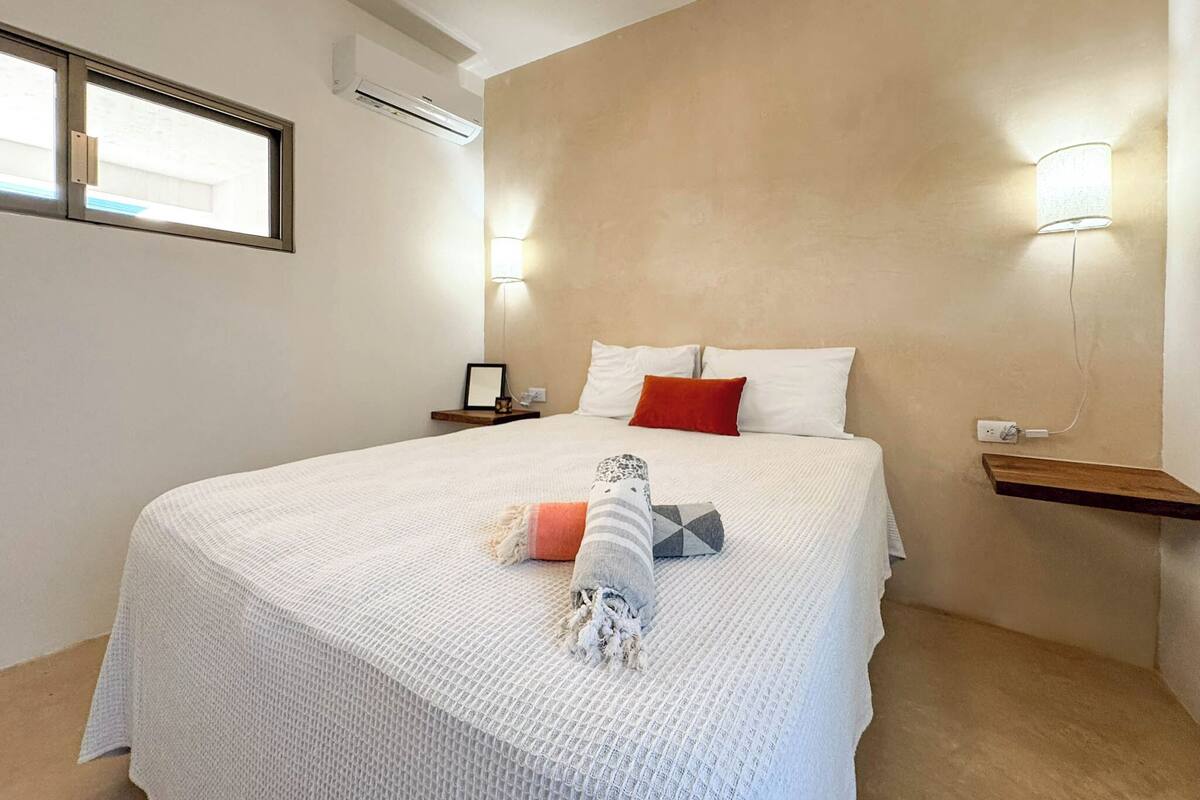 Un lit bien fait, recouvert d’une couverture blanche, avec un oreiller rouge et un plaid à rayures. Il y a deux tables de chevet avec des lampes, un climatiseur fixé au mur et une fenêtre avec des stores.