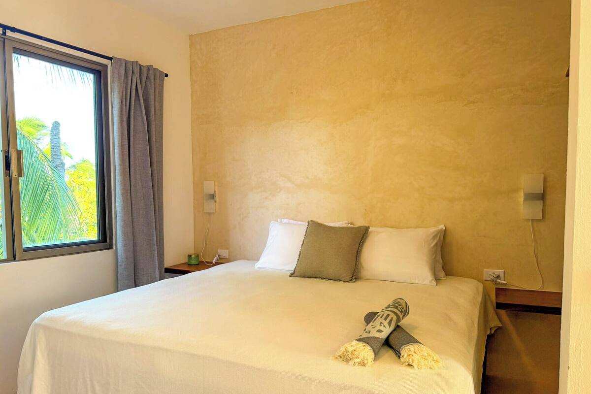 Une chambre d’hôtel avec un grand lit, une fenêtre avec des rideaux, une table de chevet et une lampe fixée au mur.