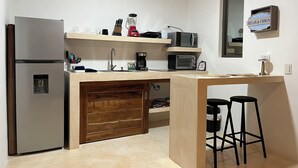 A compact kitchen with a built-in refrigerator, a microwave, a coffee maker, and a small sink. There is a wooden cabinet below the counter and two stools at the end. - Aria (El Cuyo)