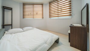 Quarto com cama, duas janelas com persianas, cômoda de madeira e espelho.