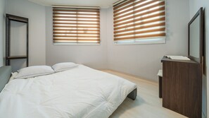 Un dormitorio con una cama, dos ventanas con persianas, un aparador de madera y un espejo.